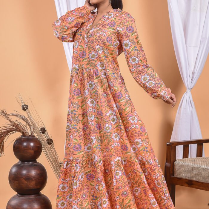 A woman in a orange floral maxi dress and with long sleeve stand in front of a light orange wall with a white drape and a table with a brass vase and flowers.