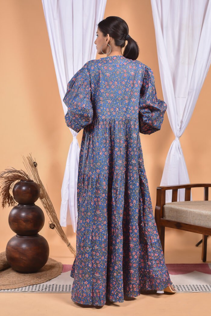 A woman in a blue floral maxi dress and with long sleeve stand in front of a light orange wall with a white drape and a table with a brass vase and flowers.