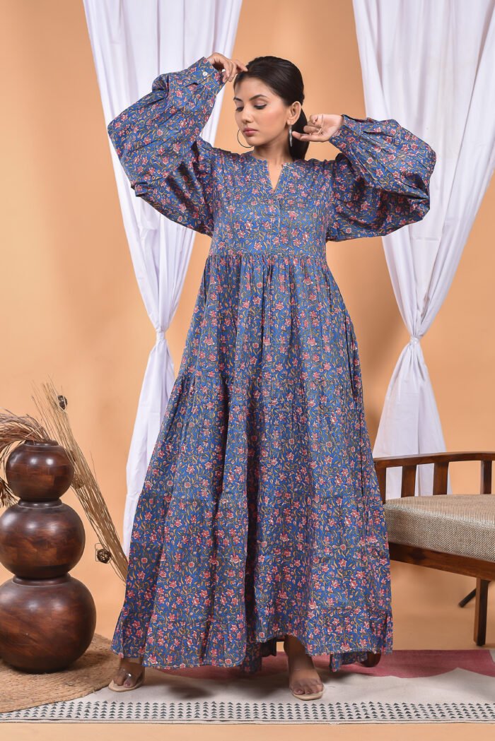 A woman in a blue floral maxi dress and with long sleeve stand in front of a light orange wall with a white drape and a table with a brass vase and flowers.