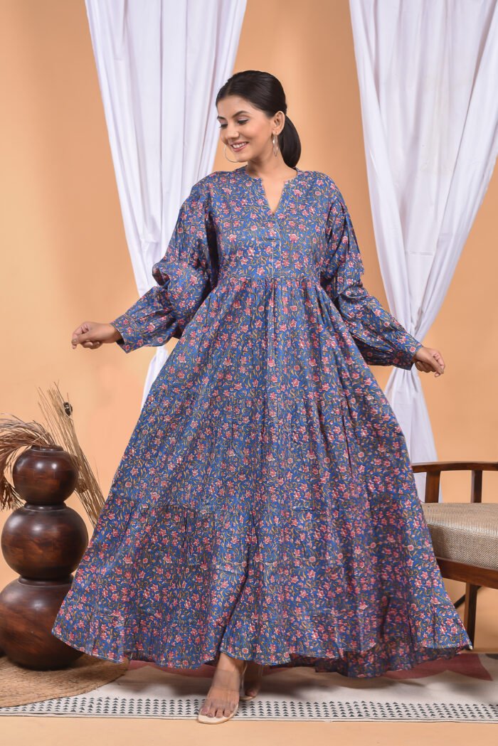 A woman in a blue floral maxi dress and with long sleeve stand in front of a light orange wall with a white drape and a table with a brass vase and flowers.