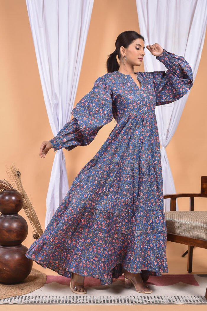 A woman in a blue floral maxi dress and with long sleeve stand in front of a light orange wall with a white drape and a table with a brass vase and flowers.