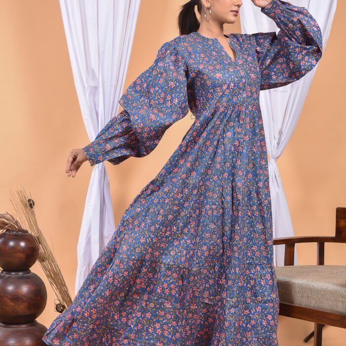 A woman in a blue floral maxi dress and with long sleeve stand in front of a light orange wall with a white drape and a table with a brass vase and flowers.