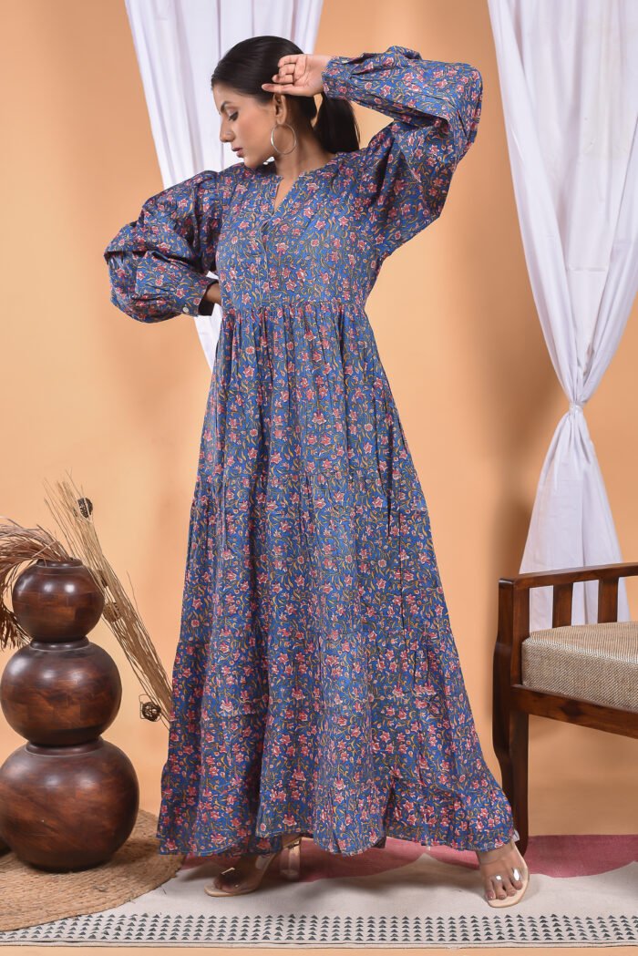 A woman in a blue floral maxi dress and with long sleeve stand in front of a light orange wall with a white drape and a table with a brass vase and flowers.