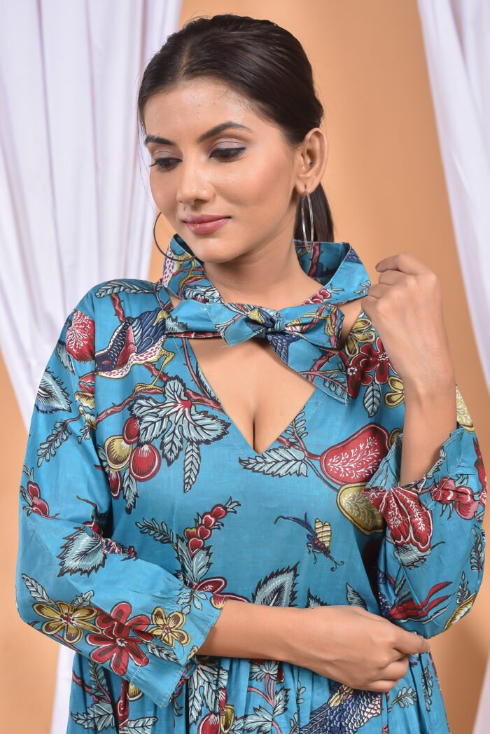 A woman in a blue floral maxi dress and with long sleeve stand in front of a light orange wall with a white drape and a table with a brass vase and flowers.