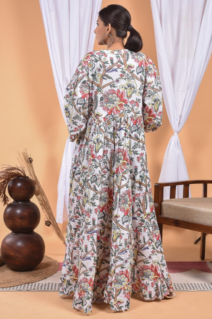 A woman in a white floral maxi dress and with long sleeve stand in front of a light orange wall with a white drape and a table with a brass vase and flowers.