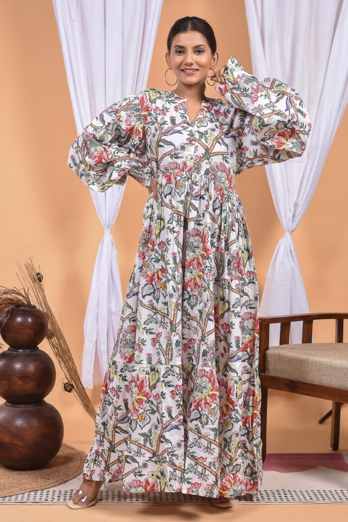 A woman in a white floral maxi dress and with long sleeve stand in front of a light orange wall with a white drape and a table with a brass vase and flowers.