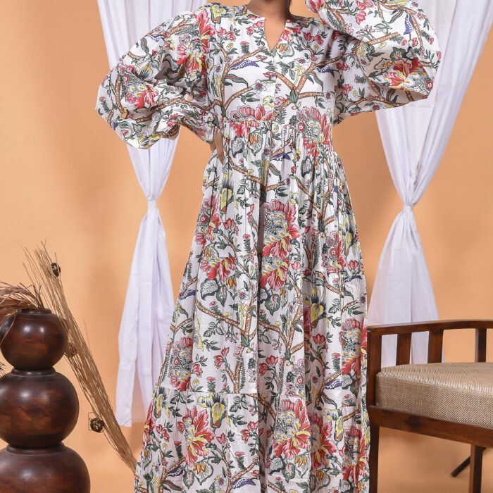 A woman in a white floral maxi dress and with long sleeve stand in front of a light orange wall with a white drape and a table with a brass vase and flowers.