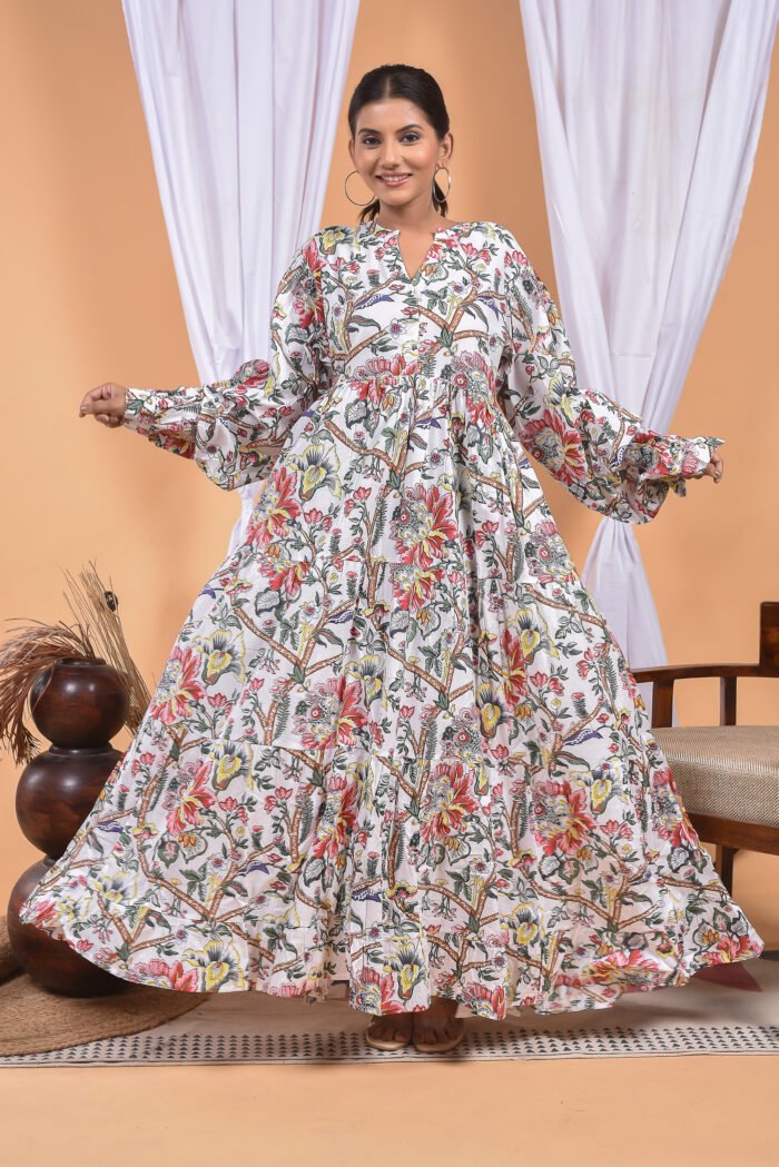 A woman in a white floral maxi dress and with long sleeve stand in front of a light orange wall with a white drape and a table with a brass vase and flowers.