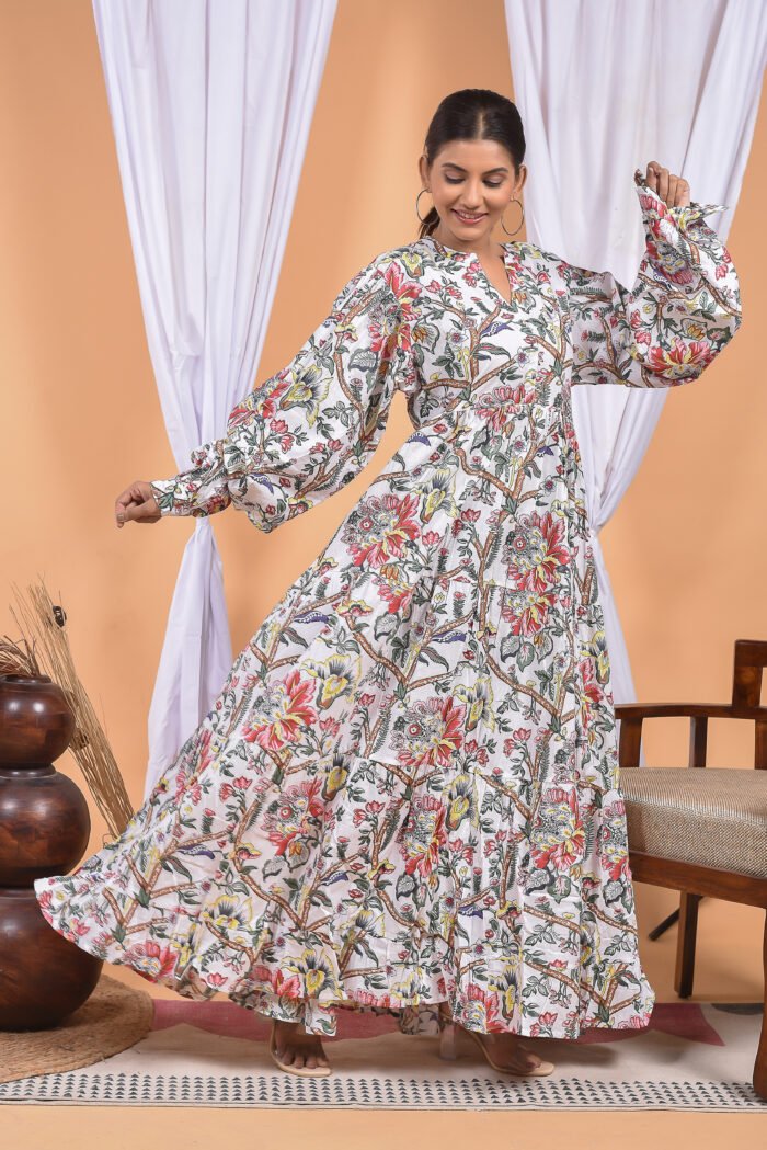 A woman in a white floral maxi dress and with long sleeve stand in front of a light orange wall with a white drape and a table with a brass vase and flowers.