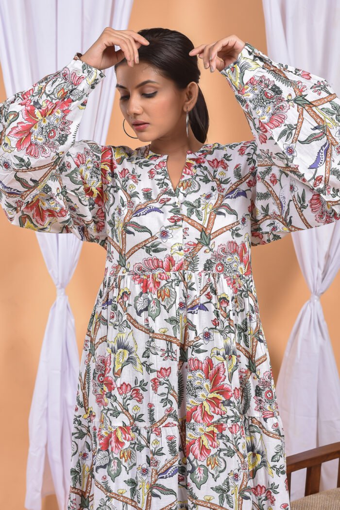 A woman in a white floral maxi dress and with long sleeve stand in front of a light orange wall with a white drape and a table with a brass vase and flowers.