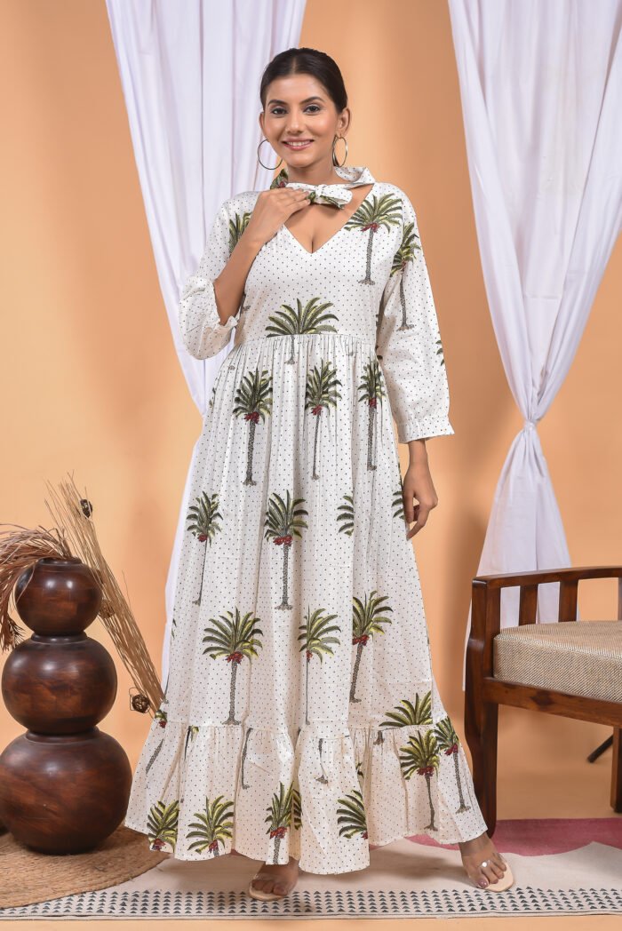 A woman in a white maxi dress with a palm tree print and long sleeve stand in front of a light orange wall with a white drape and a table with a brass vase and flowers.