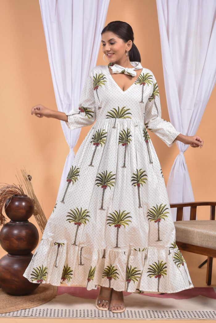 A woman in a white maxi dress with a palm tree print and long sleeve stand in front of a light orange wall with a white drape and a table with a brass vase and flowers.