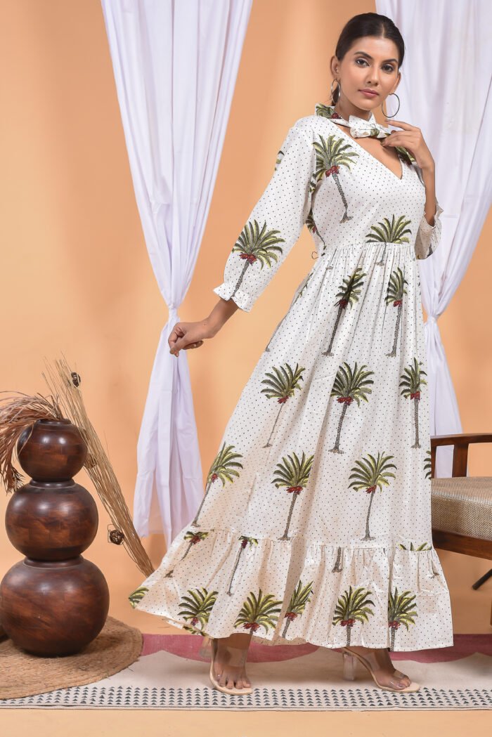 A woman in a white maxi dress with a palm tree print and long sleeve stand in front of a light orange wall with a white drape and a table with a brass vase and flowers.