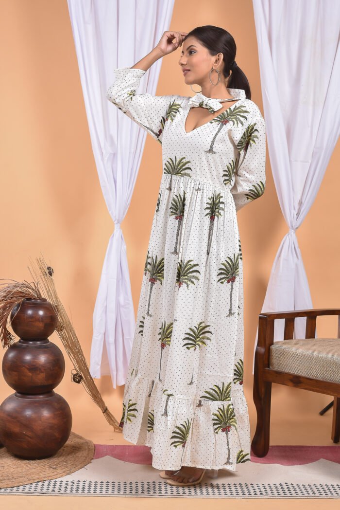 A woman in a white maxi dress with a palm tree print and long sleeve stand in front of a light orange wall with a white drape and a table with a brass vase and flowers.