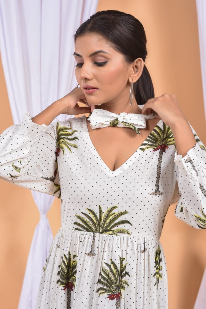 A woman in a white maxi dress with a palm tree print and long sleeve stand in front of a light orange wall with a white drape and a table with a brass vase and flowers.