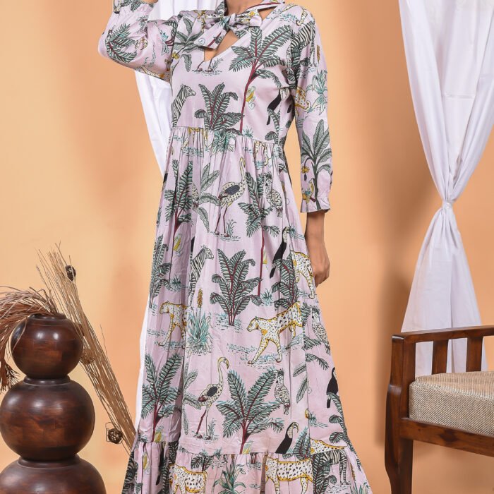 A woman in a, jungle-themed sexy light pink maxi dress with a gathered waist and halter neckline stands in front of a light orange wall with a white drape and a table with a brass vase and flowers.