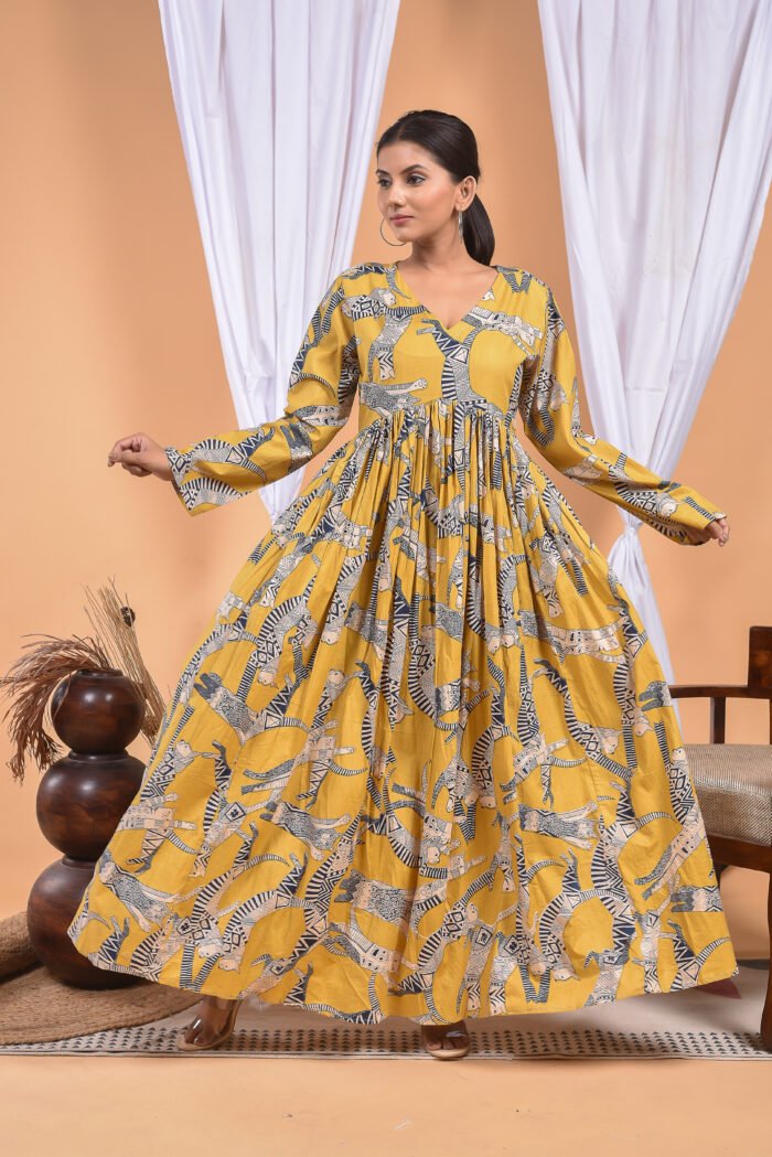 A woman in a, zebra-themed sexy yellow maxi dress with a gathered waist and halter neckline stands in front of a light orange wall with a white drape and a table with a brass vase and flowers.