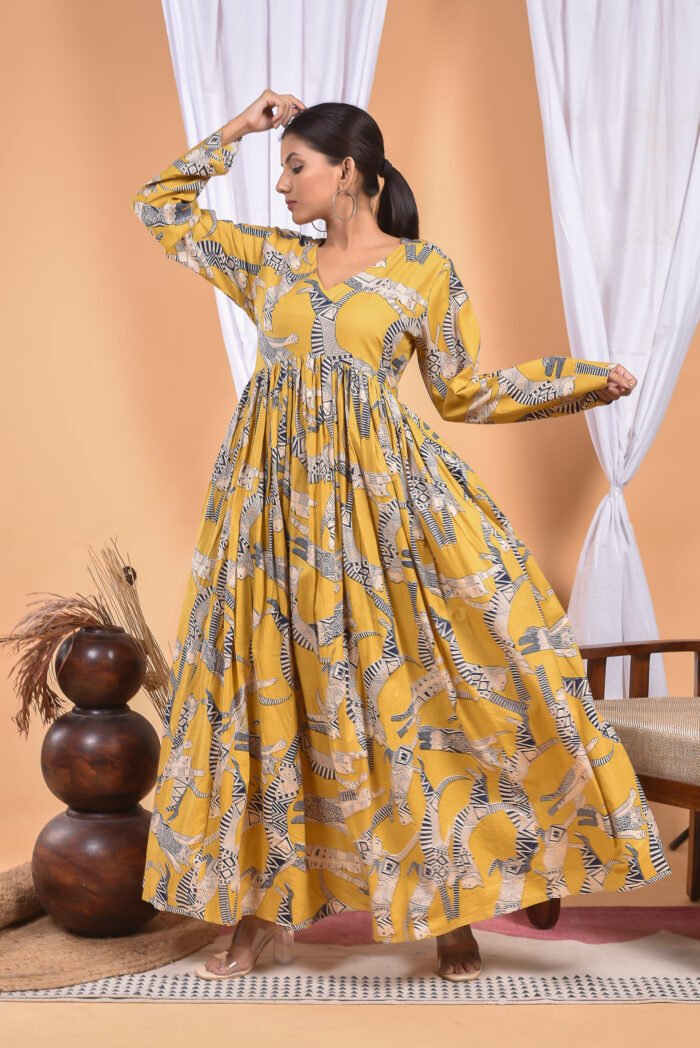 A woman in a, zebra-themed sexy yellow maxi dress with a gathered waist and halter neckline stands in front of a light orange wall with a white drape and a table with a brass vase and flowers.