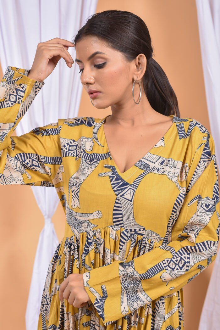 A woman in a, zebra-themed sexy yellow maxi dress with a gathered waist and halter neckline stands in front of a light orange wall with a white drape and a table with a brass vase and flowers.