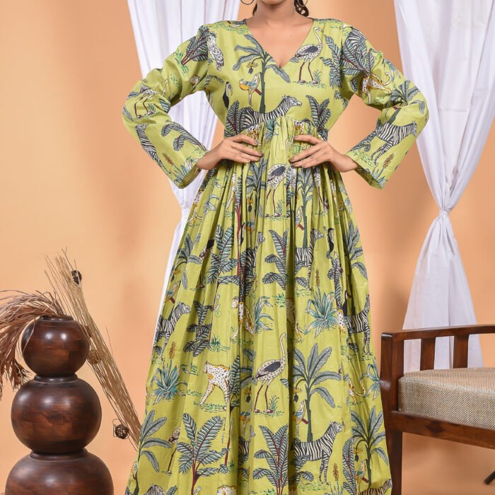 A woman in a, jungle-themed green boho maxi dress with sleeves and with a gathered waist and halter neckline stands in front of a light orange wall with a white drape and a table with a brass vase and flowers.