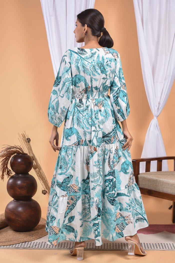 A woman in a, floral-themed blue floral maxi dress with a gathered waist and halter neckline stands in front of a light orange wall with a white drape and a table with a brass vase and flowers.