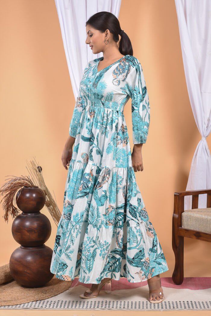 A woman in a, floral-themed blue floral maxi dress with a gathered waist and halter neckline stands in front of a light orange wall with a white drape and a table with a brass vase and flowers.