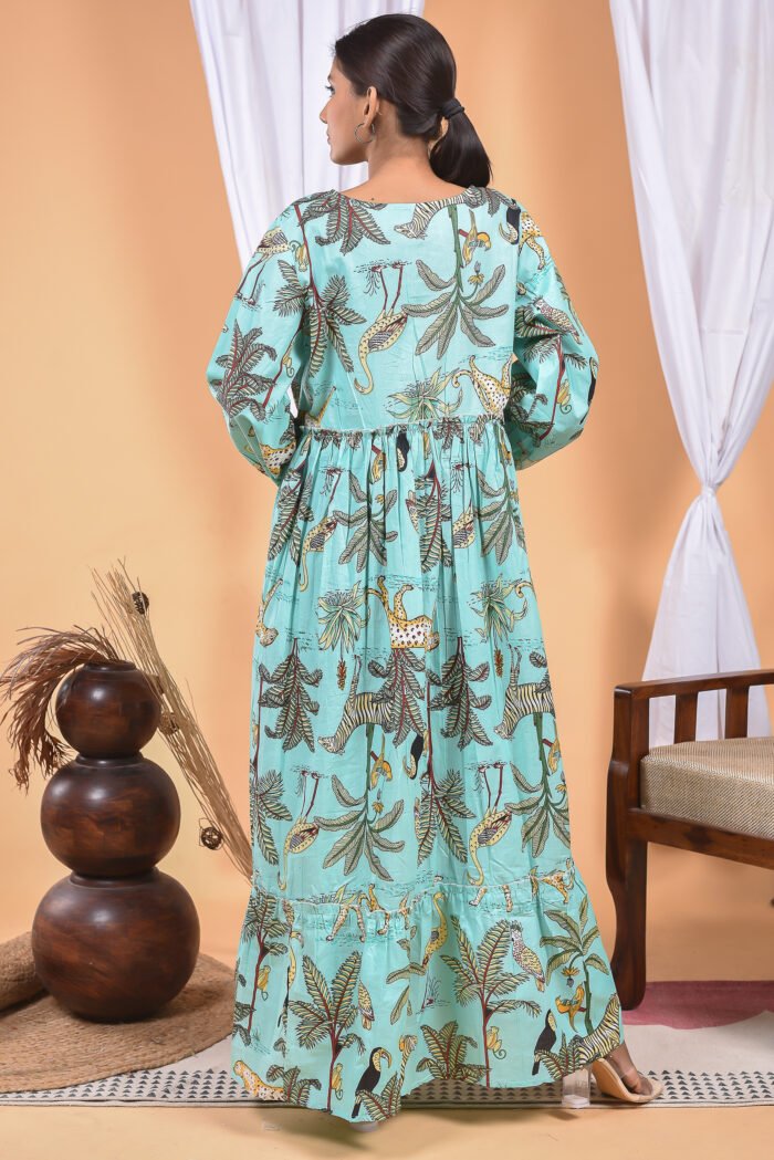 A woman in a, jungle-themed blue maxi dress with a gathered waist and halter neckline stands in front of a light orange wall with a white drape and a table with a brass vase and flowers.