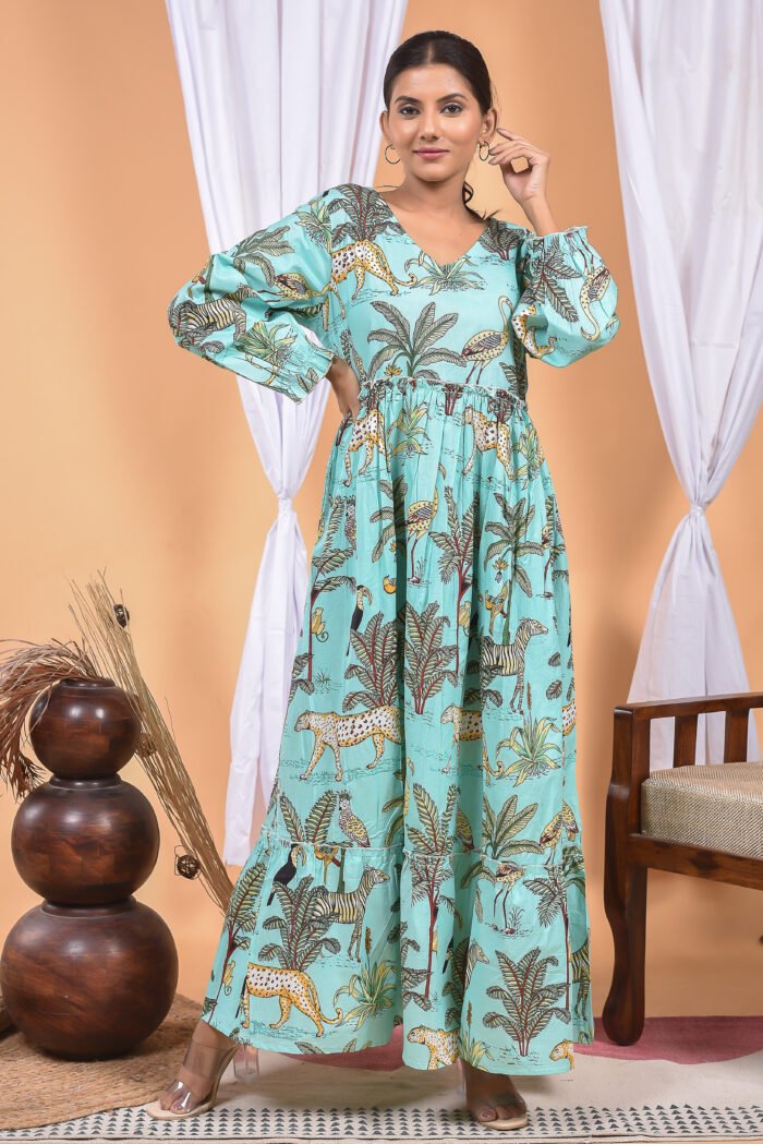 A woman in a, jungle-themed blue maxi dress with a gathered waist and halter neckline stands in front of a light orange wall with a white drape and a table with a brass vase and flowers.