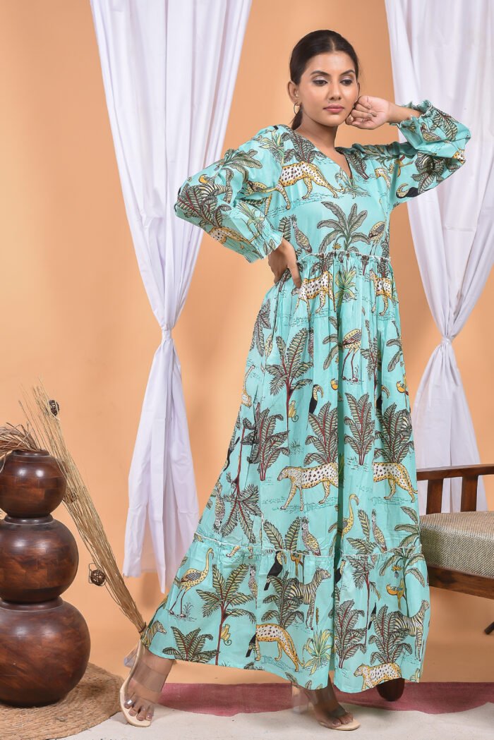 A woman in a, jungle-themed blue maxi dress with a gathered waist and halter neckline stands in front of a light orange wall with a white drape and a table with a brass vase and flowers.