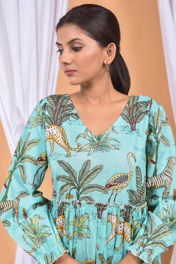 A woman in a, jungle-themed blue maxi dress with a gathered waist and halter neckline stands in front of a light orange wall with a white drape and a table with a brass vase and flowers.