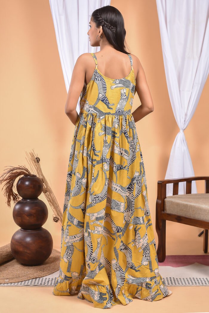 A woman in a, zebra-themed sexy yellow maxi dress with a gathered waist and halter neckline stands in front of a light orange wall with a white drape and a table with a brass vase and flowers.