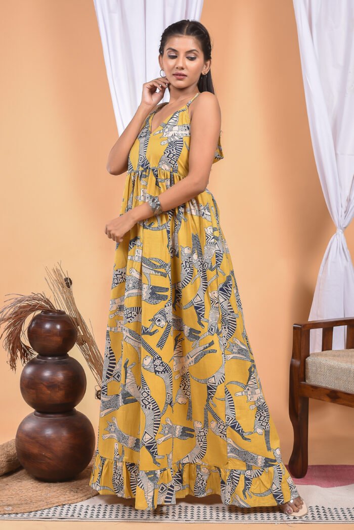 A woman in a, zebra-themed sexy yellow maxi dress with a gathered waist and halter neckline stands in front of a light orange wall with a white drape and a table with a brass vase and flowers.