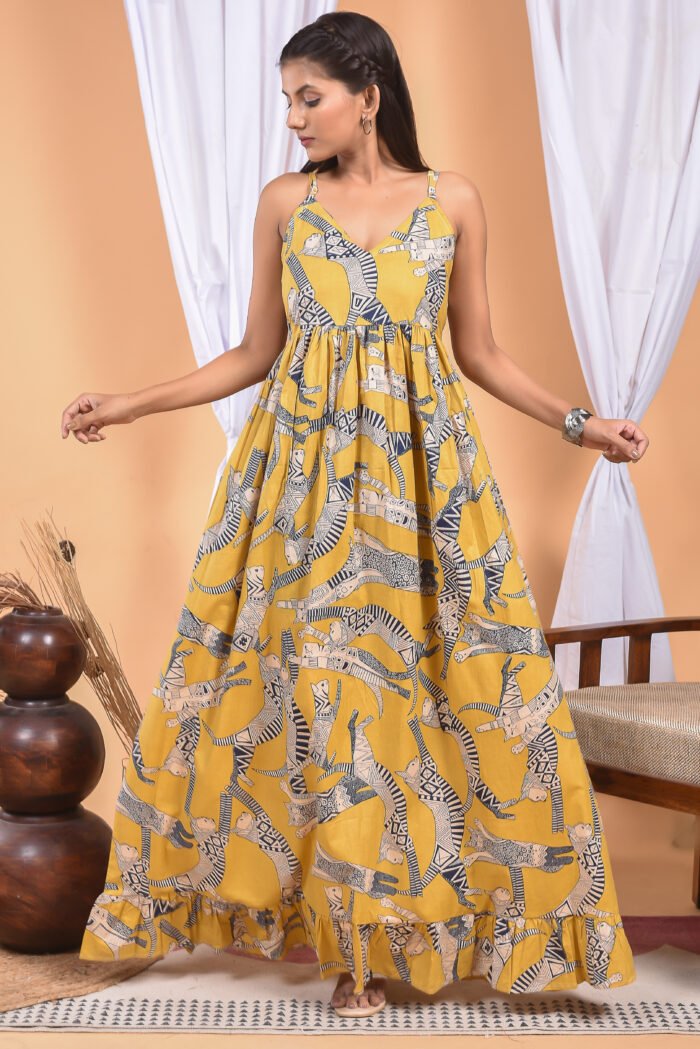 A woman in a, zebra-themed sexy yellow maxi dress with a gathered waist and halter neckline stands in front of a light orange wall with a white drape and a table with a brass vase and flowers.