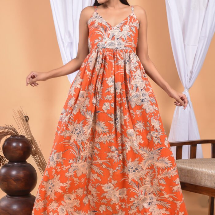 A woman in a, floral-themed sexy orange maxi dress with a gathered waist and halter neckline stands in front of a light orange wall with a white drape and a table with a brass vase and flowers.