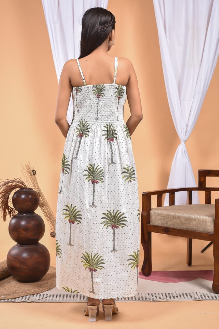 A woman in a white maxi dress with a palm tree print and halter neckline stand in front of a light orange wall with a white drape and a table with a brass vase and flowers.