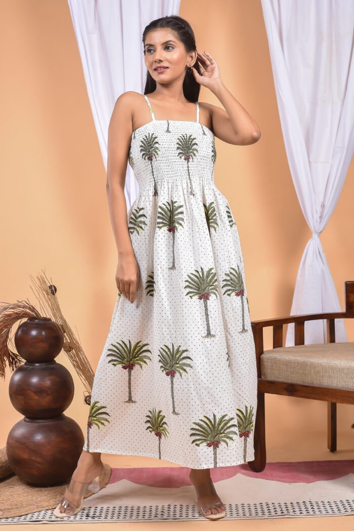 A woman in a white maxi dress with a palm tree print and halter neckline stand in front of a light orange wall with a white drape and a table with a brass vase and flowers.