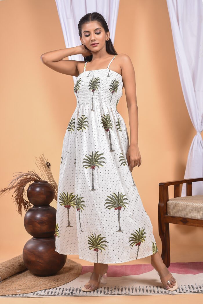 A woman in a white maxi dress with a palm tree print and halter neckline stand in front of a light orange wall with a white drape and a table with a brass vase and flowers.