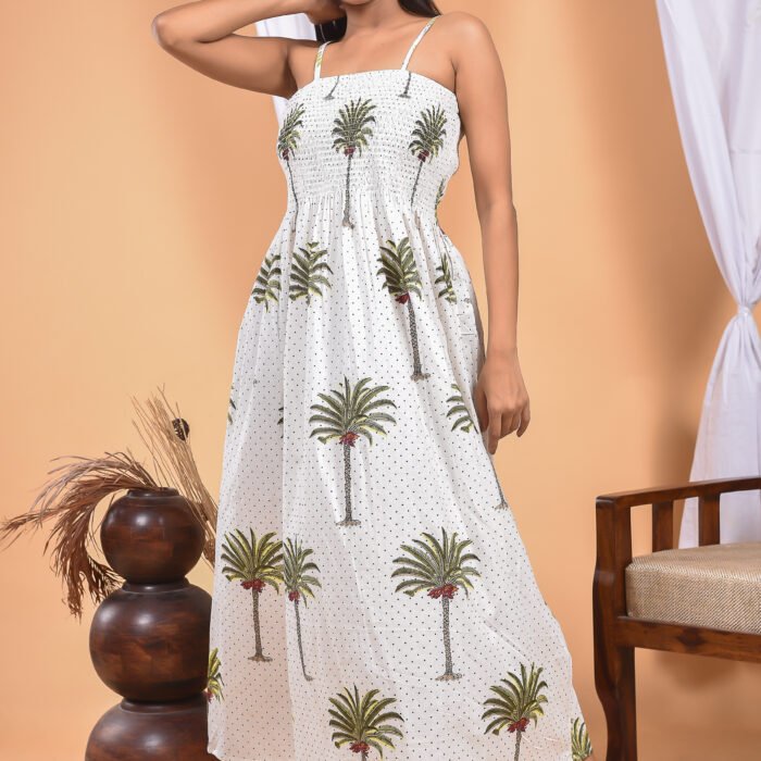 A woman in a white maxi dress with a palm tree print and halter neckline stand in front of a light orange wall with a white drape and a table with a brass vase and flowers.