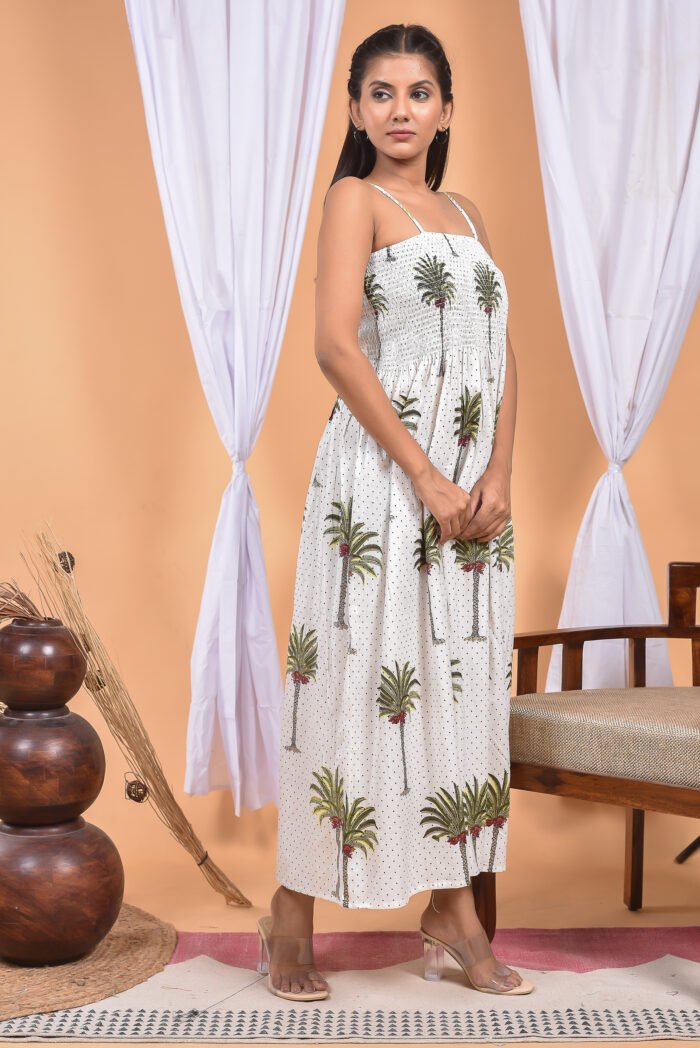 A woman in a white maxi dress with a palm tree print and halter neckline stand in front of a light orange wall with a white drape and a table with a brass vase and flowers.