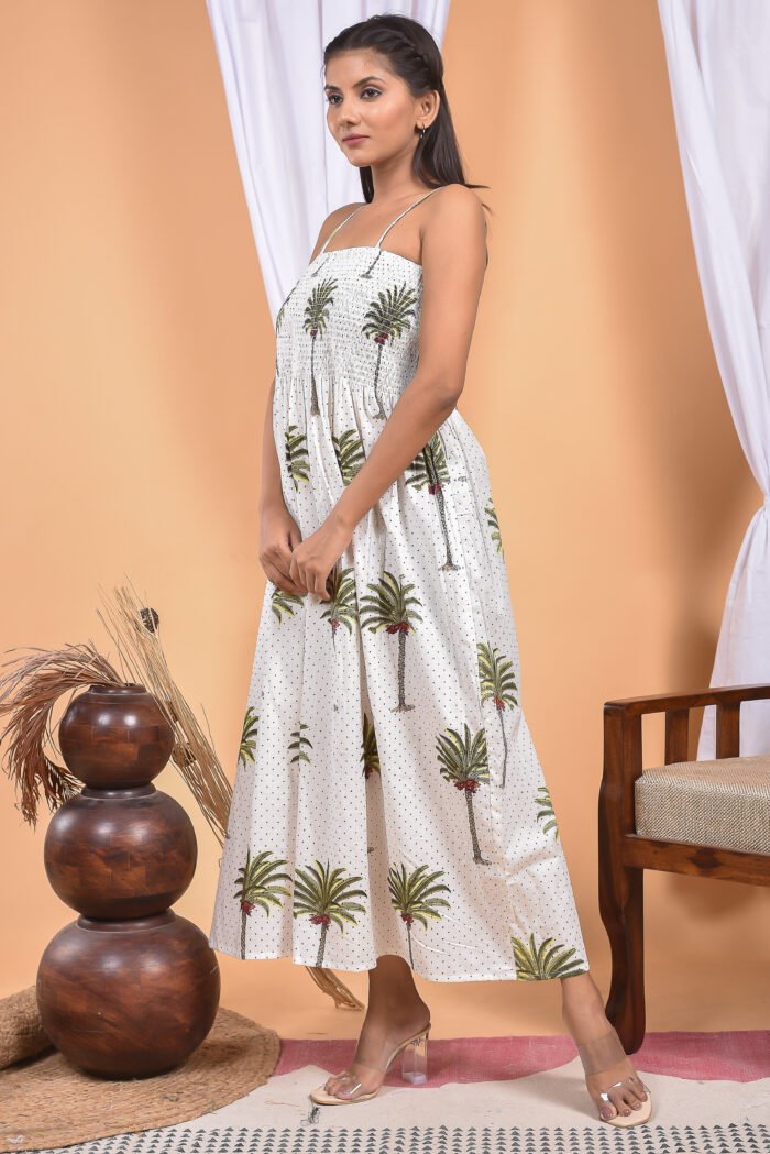 A woman in a white maxi dress with a palm tree print and halter neckline stand in front of a light orange wall with a white drape and a table with a brass vase and flowers.