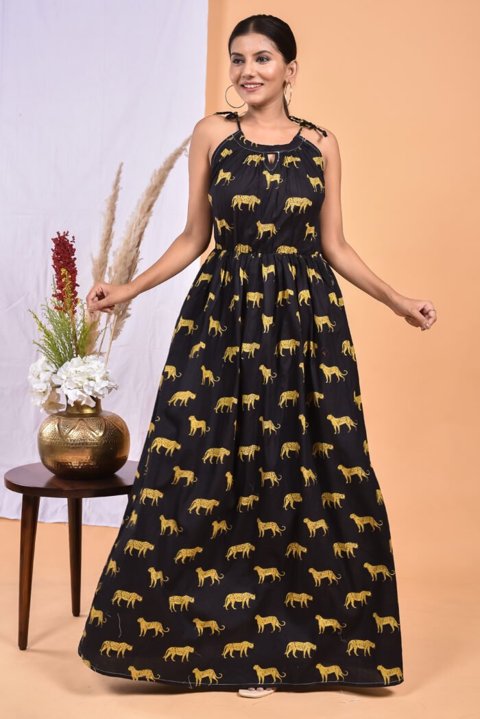 A woman in a, black leopard maxi dress with a gathered waist and halter neckline stands in front of a light orange wall with a white drape and a table with a brass vase and flowers.