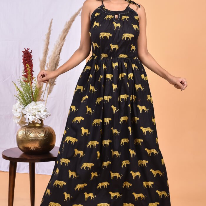 A woman in a, black leopard maxi dress with a gathered waist and halter neckline stands in front of a light orange wall with a white drape and a table with a brass vase and flowers.