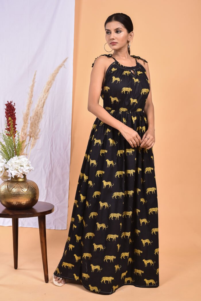 A woman in a, black leopard maxi dress with a gathered waist and halter neckline stands in front of a light orange wall with a white drape and a table with a brass vase and flowers.