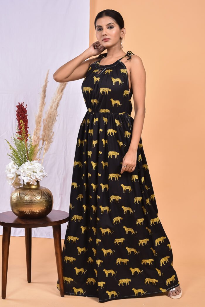 A woman in a, black leopard maxi dress with a gathered waist and halter neckline stands in front of a light orange wall with a white drape and a table with a brass vase and flowers.