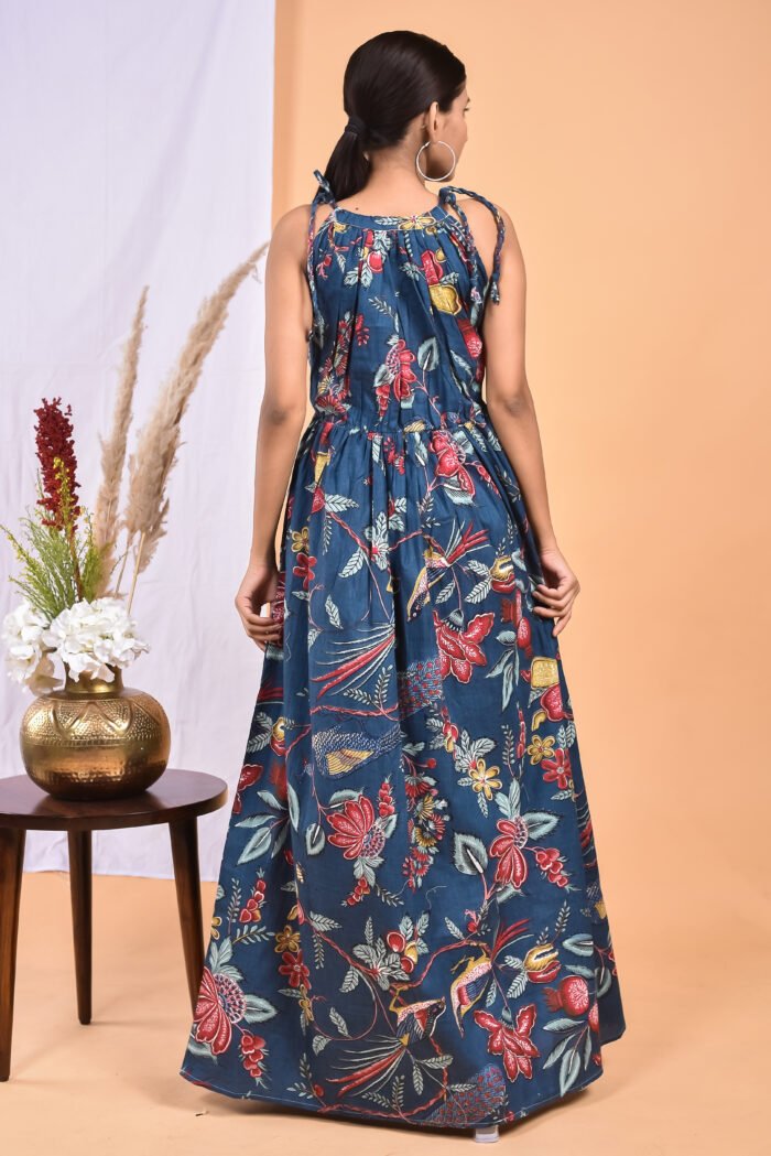 A woman in a blue floral maxi dress with a gathered waist and halter neckline stands in front of a light orange wall with a white drape and a table with a brass vase and flowers.