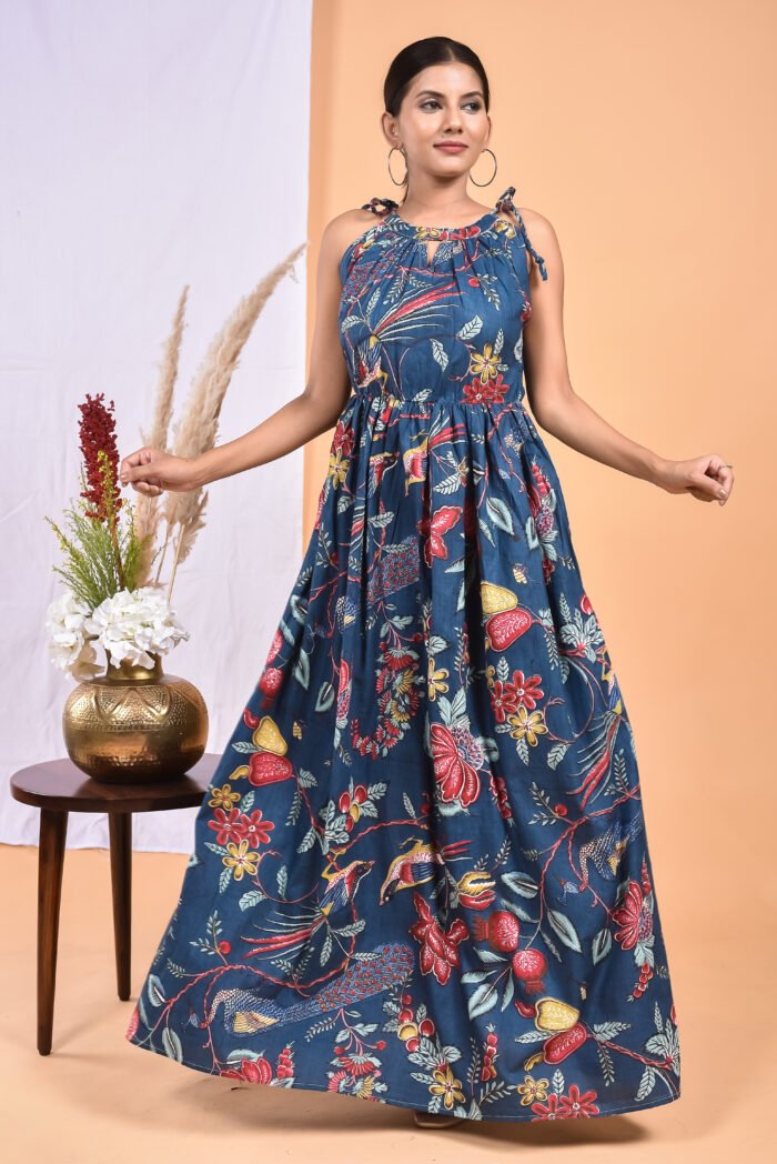 A woman in a blue floral maxi dress with a gathered waist and halter neckline stands in front of a light orange wall with a white drape and a table with a brass vase and flowers.