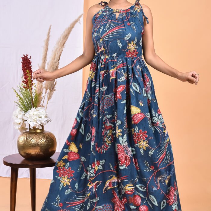 A woman in a blue floral maxi dress with a gathered waist and halter neckline stands in front of a light orange wall with a white drape and a table with a brass vase and flowers.