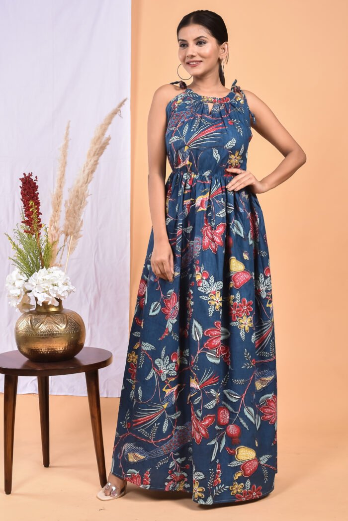 A woman in a blue floral maxi dress with a gathered waist and halter neckline stands in front of a light orange wall with a white drape and a table with a brass vase and flowers.