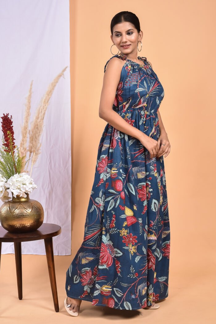 A woman in a blue floral maxi dress with a gathered waist and halter neckline stands in front of a light orange wall with a white drape and a table with a brass vase and flowers.