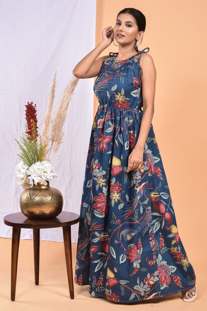 A woman in a blue floral maxi dress with a gathered waist and halter neckline stands in front of a light orange wall with a white drape and a table with a brass vase and flowers.